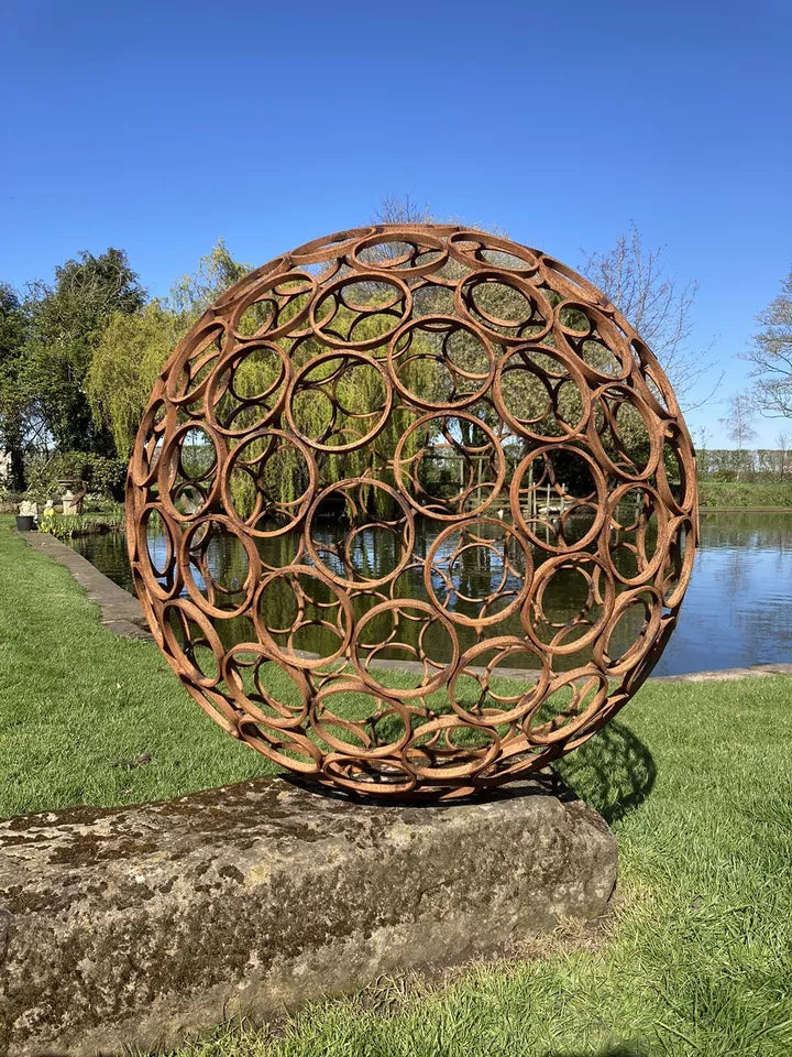 Rustic steel garden sphere sculpture with circular pattern, displayed on a stone base by a pond, enhancing outdoor landscape aesthetics.