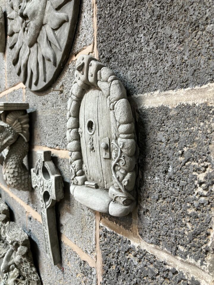Stone Fairy Door Plaque displayed among other garden ornaments on a textured wall, showcasing its antique grey limestone finish and fairy-tale charm.