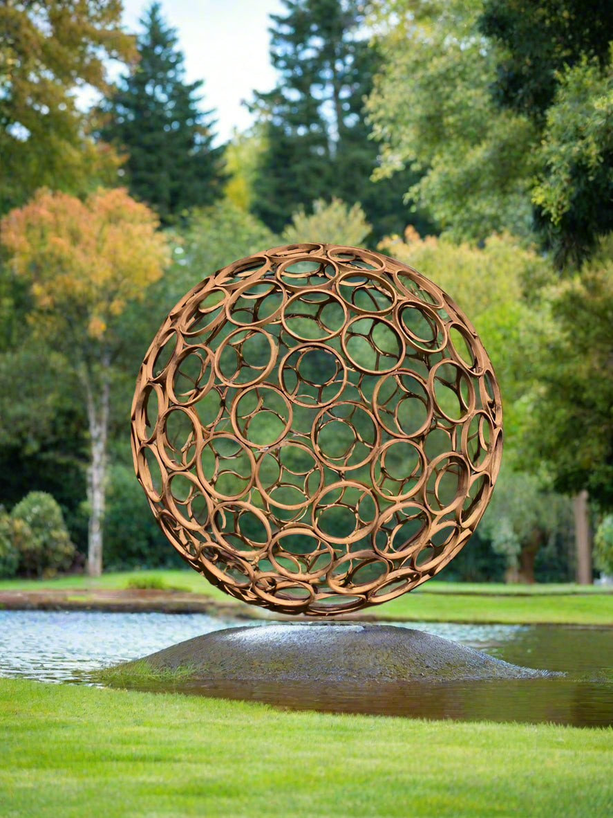 Weathered steel garden sphere sculpture with open ring design, positioned on a mound near water, adding rustic elegance to outdoor spaces.