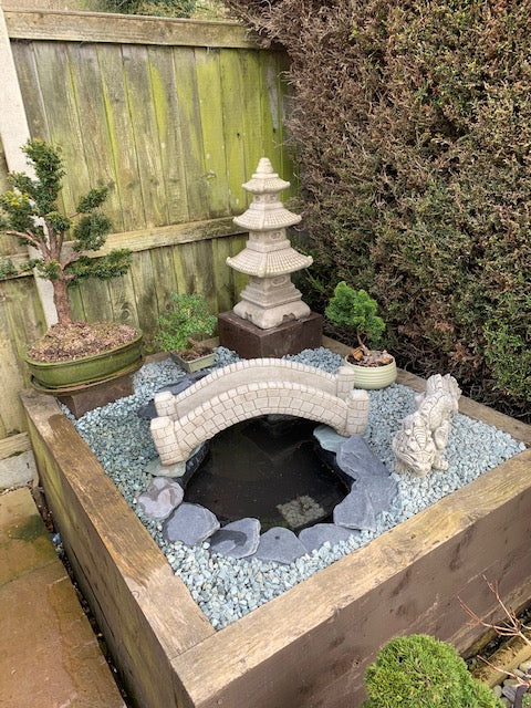 Stone pond bridge garden ornament with antique finish placed over a small pond, surrounded by bonsai trees and Asian-style decor.