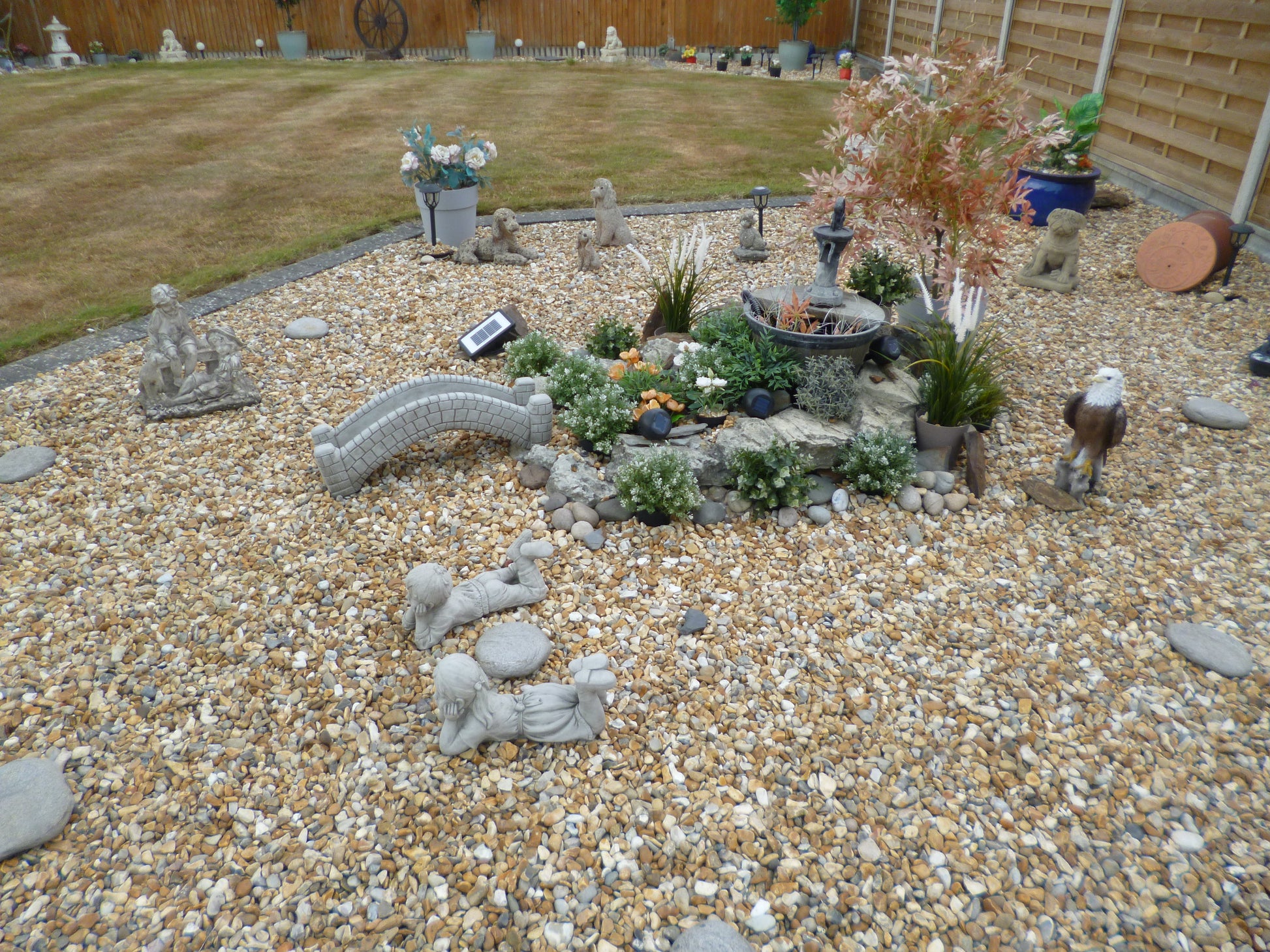 Garden ornament - stone pond bridge with antique finish in a landscaped pebble garden, surrounded by plants, statues, and solar lighting.